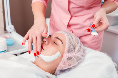 Beautician apply facial cream on young woman face skin. Cosmetic procedure in spa salon. Mask for facial skin care.の写真素材