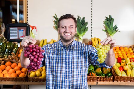 Portrait of smiling man in shirt selling ripe grapes in shop. Boxes with fruits and vegetables on the background.の写真素材
