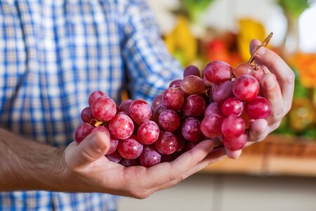 Portrait of happy man in shirt showing a bunch of grapes in supermarket. Boxes with fruits and vegetables on the background.の写真素材