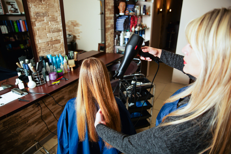 Professional woman hairdresser making hairstyle using hair dryer for young female in beauty salon.の写真素材