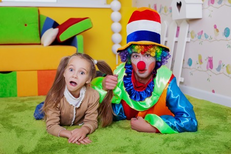 Smiling child and the clown lie on a carpet in the nursery. Colourful sofa, nesting box and fence on the background.の写真素材