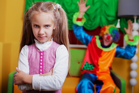 Upset little girl standing in the playroom.  Funny clown on the background.の写真素材
