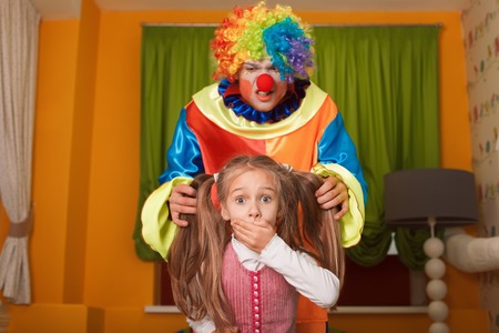 Little girl was frightened of the clown who has crept behind. Kindergarten with colorful couch on the background.の写真素材