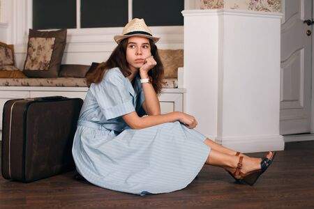 Vintage travelling concept with a young woman in hat sitting on the floor nearby her suitcase in the living room. Sofa and window with curtains on the background.の写真素材
