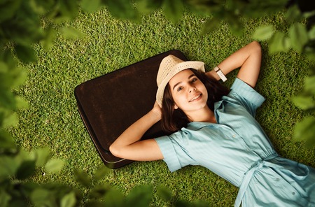 Young woman in light blue dress and white hat put her head on suitcase and dreams of a future journey. Girl lies on green grass. Travel waiting concept.の写真素材
