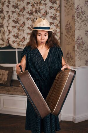 Young female in black dress holding a half-open suitcase in her hands. Vintage style. Sofa and window with curtains on the background.の写真素材