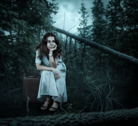 Young woman in white dress sitting on her suitcase in the forest. Vintage travel concept.の写真素材