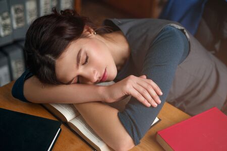 Young girl fell asleep while reading a book in college library. Bookshelf on the background. Student...