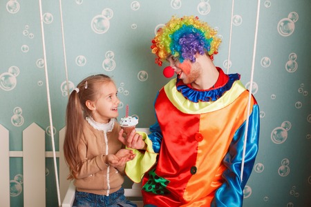 Joyful little girl with happy clown sitting on a swing, he invites her to eat birthday cakeの写真素材