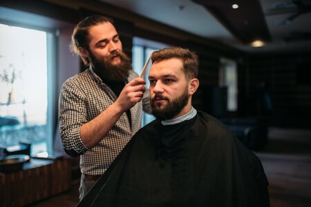 Barber combing hair of the client in salon cape