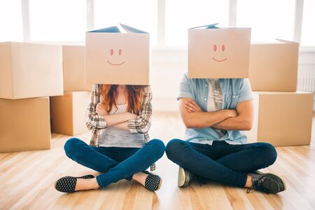 Playful couple with cardboard boxes on their heads sitting on the floor, moving to new home, housewarming. Man and woman have a funの写真素材