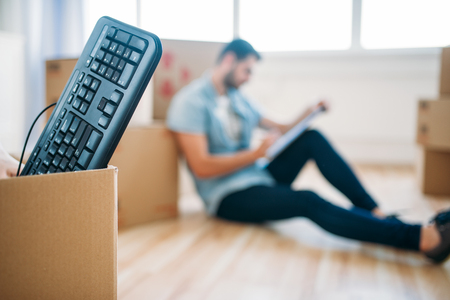 Box with keyboard, male person with notebook among cardboard boxes on background, housewarming....