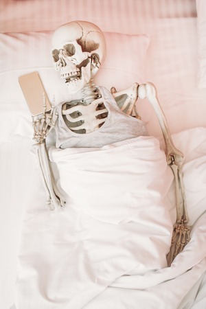Human skeleton with phone in hand lies in bedの写真素材
