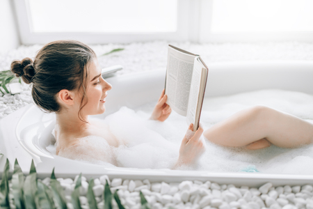 Woman lying in bath with foam and reads magazineの写真素材
