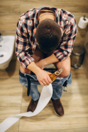 Drunk man with bottle of beer sitting on toiletの写真素材