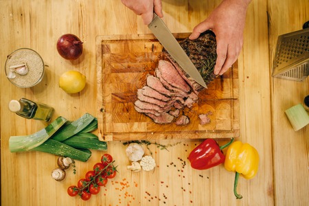 Male chef hands with knife cuts roasted meat on slices, top view. Man preparing beef with vegetables on countertopの写真素材