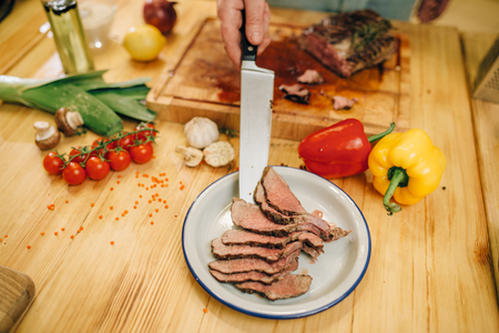 Male chef with knife puts roasted meat slices into the plate, kitchen on background. Man preparing beef with vegetables on countertopの写真素材