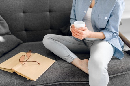 Young lady with book and cup of coffee poses on cozy black couch, living room in white tones on background. Attractive female person with magazine sitting on sofa at homeの写真素材