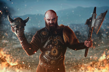 Viking holds axe and human skull, battle in fire