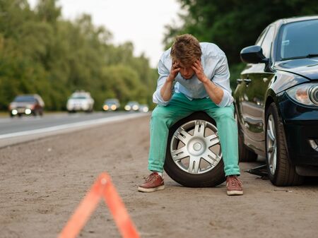 Car breakdown, tired man sitting on spare tyre. Broken automobile or problem with vehicle, trouble with punctured auto tire on highway, emergency stop signの写真素材