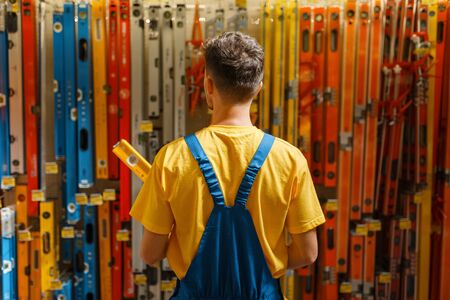 Male builder with building level in hardware store. Constructor in uniform look at the goods in diy...