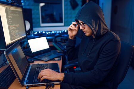 Hacker in hood at his workplace with laptop and desktop PC, website or corporate hacking, darknet...