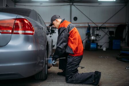 Mechanic in uniform unscrews the wheel in tire service. Man repairs car tyre in garage, automobile inspection in workshopの写真素材