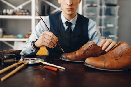 Bootmaker with brush tints shoes, footwear repair service. Craftsman skill, shoemaking workshop,...