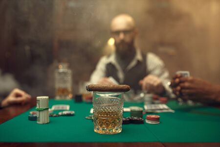 Whiskey and cigar on gaming table with green cloth, bearded poker player on background, casino. Games of chance. Man leisures in gambling houseの写真素材