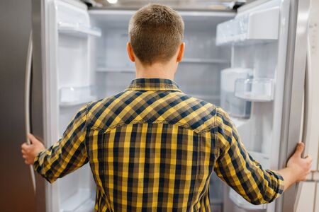 Young man at the opened refrigerator in electronics store. Male person buying home electrical...