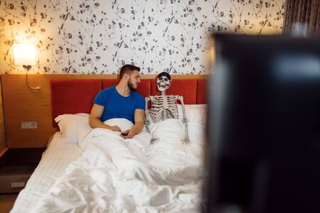 Man and human skeleton watching TV in the bedの写真素材