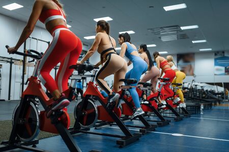 Group of women doing exercise on a stationary bike in gym, back view. People on fitness workout in sport club, athletic girls in sportswear on training indoorsの写真素材