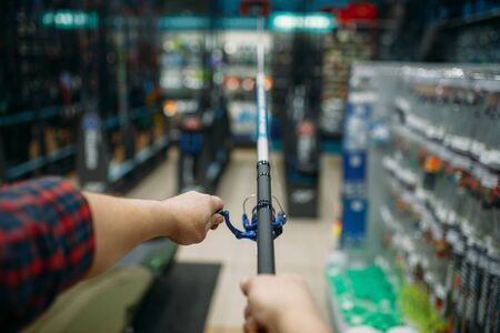 Male angler with rod in fishing shop, first-person view. Equipment and tools for fish catching and hunting, accessory choice on showcase in store, spinnings and telescopes assortmentの写真素材