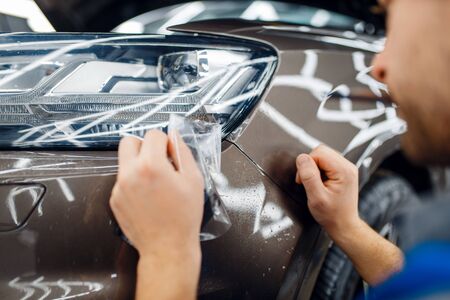 Male worker installs transparent protection film on car headlight. Installation of coating that protects the paint of automobile from scratches. New vehicle in garage, autotuningの写真素材