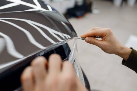 Male worker installs transparent protection film on car hood. Installation of coating that protects the paint of automobile from scratches. New vehicle in garageの写真素材