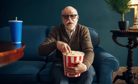 Elderly man with popcorn watching TV on couchの写真素材