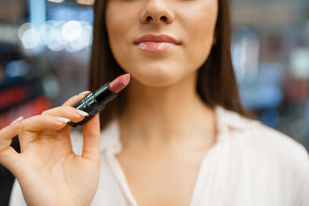 Woman holds lipstick at her face in cosmetics store. Buyer in luxury beauty shop salon, female...