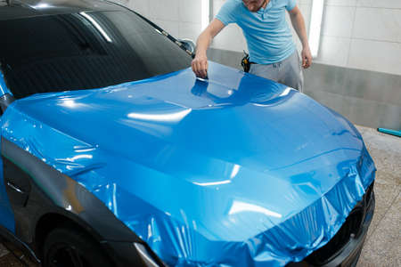 Car wrapping, man with squeegee installs protective vinyl foil or film on hood. Worker makes auto detailing. Automobile paint protection coating, professional tuningの写真素材