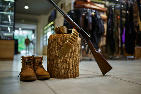 Rifle and hunting boots at the stump in gun store, nobody. Weapon shop interior on background, ammunition assortment, firearms choice, shooting hobby and lifestyle, self protection and securityの写真素材