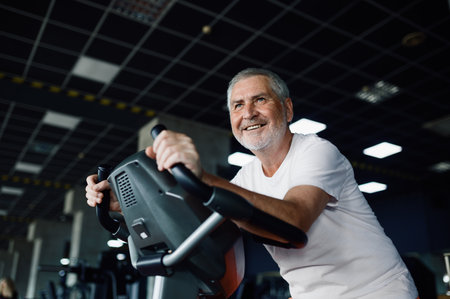 Elderly man poses on exercise bike in gymの写真素材
