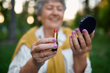 Pretty elderly woman holds lipstick in summer parkの写真素材