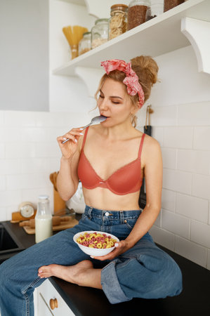Young slim woman in lingerie bra and jeans eating flakes with milk