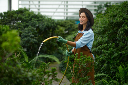 Young woman gardener watering green plants from hose in greenhouseの写真素材