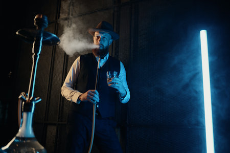 Front view of resting man smoking hookah at home workplace with neon lighting