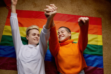 Happy young lesbian couple lying above lgtb rainbow flag with raised hands upの写真素材