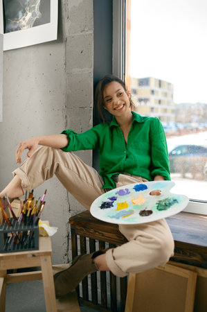 Happy smiling woman sitting on windowsill holding paint palette in handsの写真素材