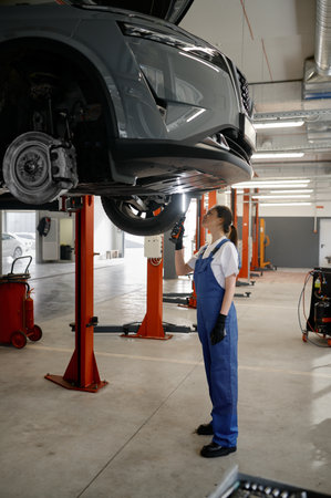 Woman mechanic examining car suspended on car lift using flashlightの写真素材