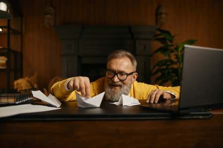 Happy excited mature grey-haired man playing with paper boat at workplace