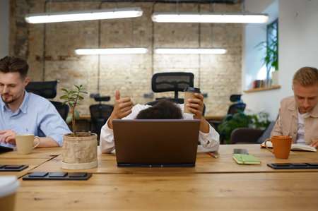 Woman office worker hiding head behind laptop gesturing thumbs upの写真素材
