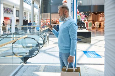 Rich senior man booking taxi online standing with shopping bags in supermarketの写真素材